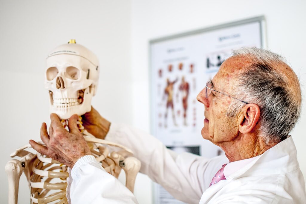 Profesional de la salud examinando un modelo anatómico de esqueleto humano en un consultorio médico. La imagen destaca la importancia de la educación en anatomía y los métodos para prevenir la pérdida ósea, con un póster de referencia anatómica en el fondo.
