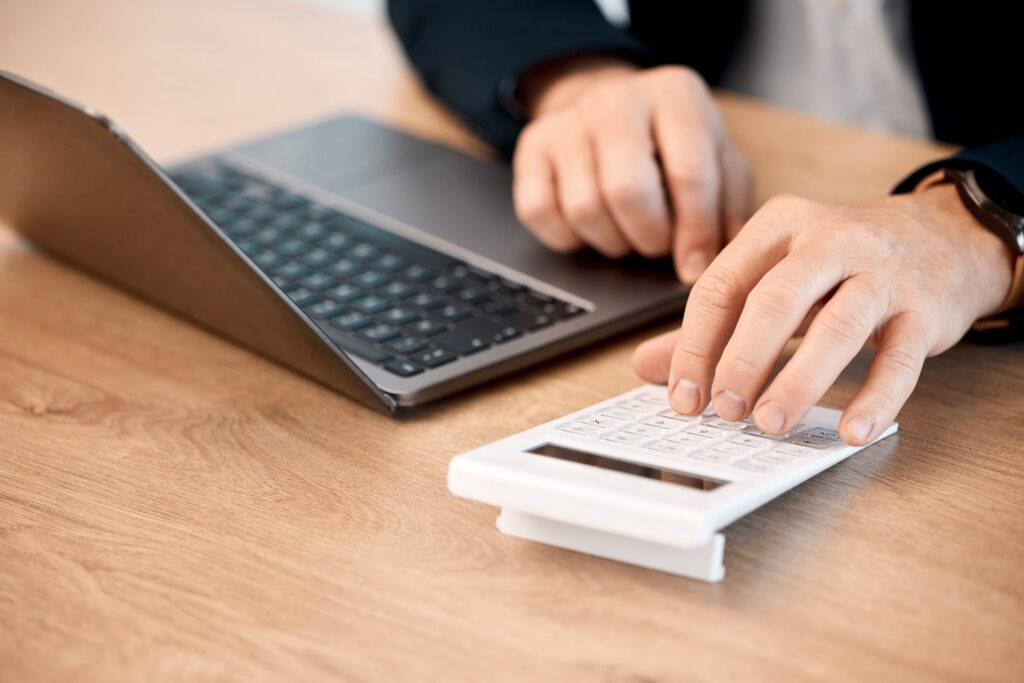 Manos utilizando una calculadora junto a un portátil sobre una mesa, representando el análisis financiero y la estimación del coste de los tratamientos adicionales.