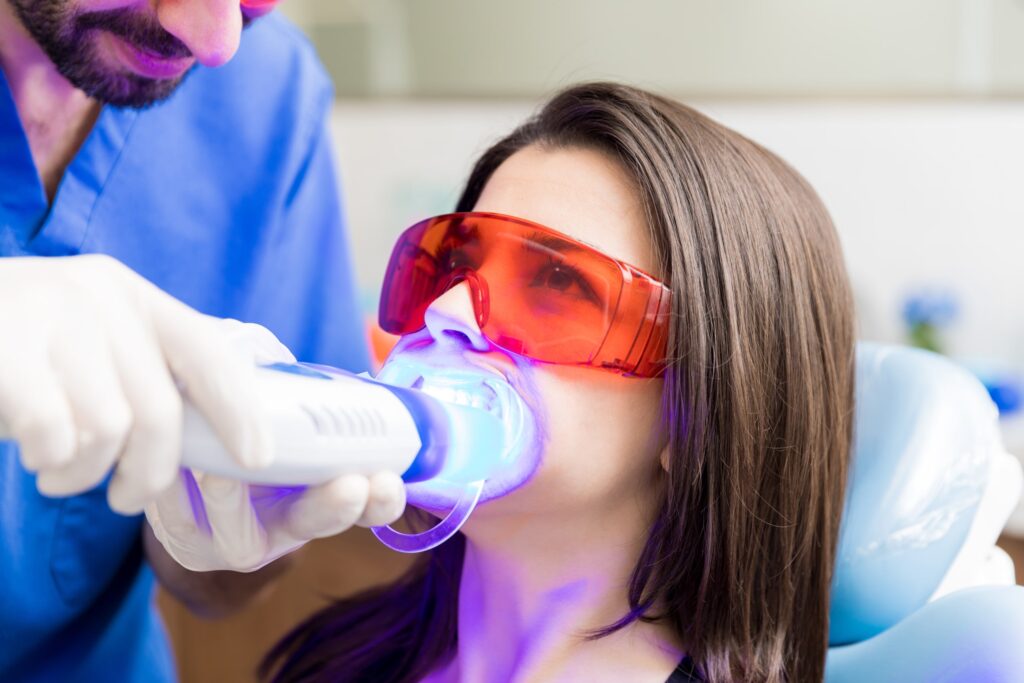 Mujer recibiendo un tratamiento de blanqueamiento dental con luz LED en una clínica odontológica, usando gafas de protección y siendo atendida por un dentista. Esta imagen ilustra los efectos del blanqueamiento dental y la importancia de los cuidados posteriores para mantener resultados óptimos.