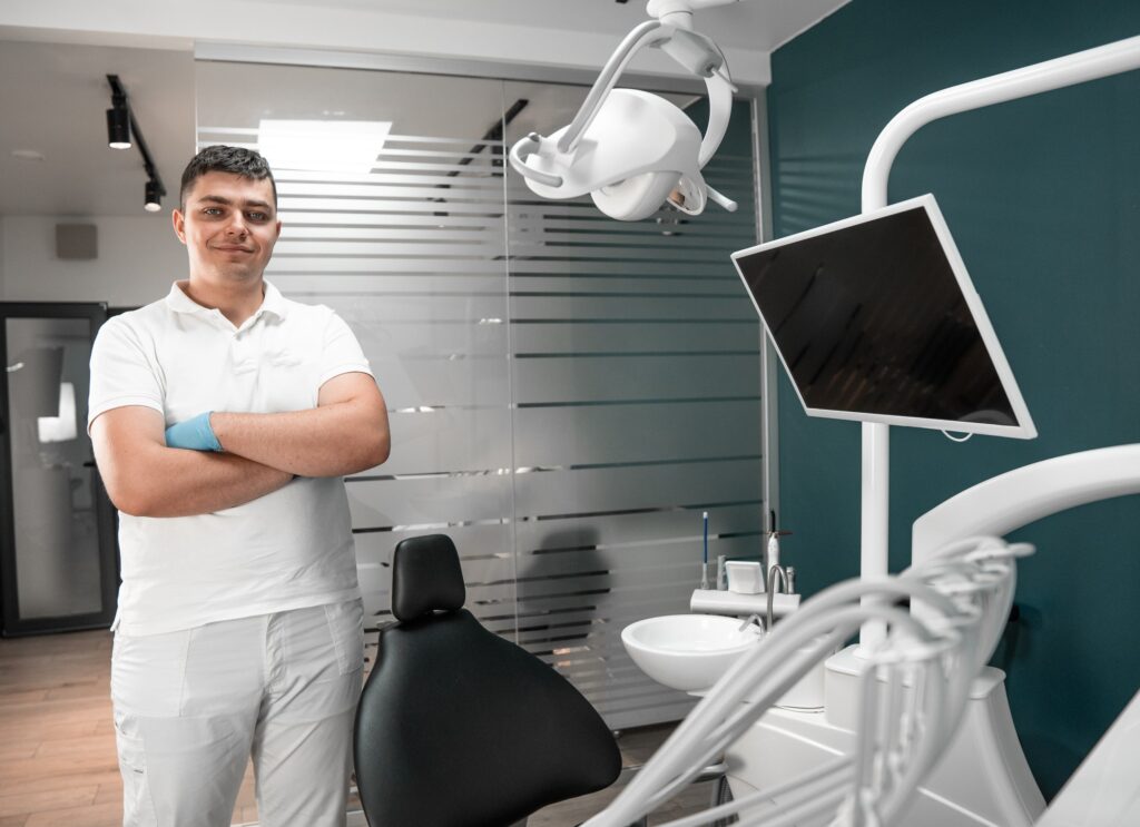 Un dentista en su consultorio, vestido con uniforme blanco y guantes, de pie junto a una silla dental y equipo especializado. La imagen representa un ambiente profesional para destacar las recomendaciones de limpieza dental.