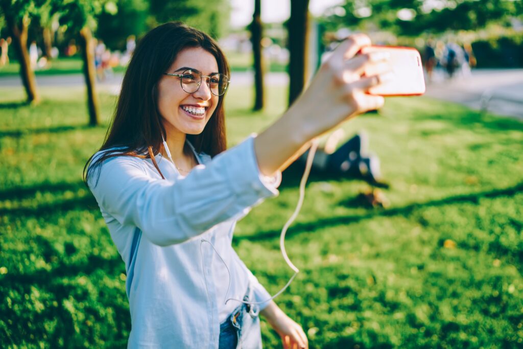Mujer joven con dientes blancos y brillantes sonríe mientras se toma una selfie en un parque, ilustrando cómo factores como la alimentación, la higiene y los hábitos pueden influir en la duración del blanqueamiento dental. Palabra clave: Factores que influyen en la duración del blanqueamiento.