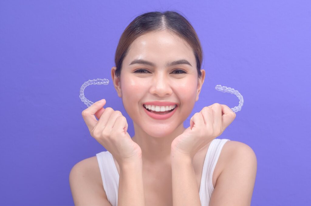 Mujer sostiene dos alineadores transparentes Invisalign frente a un fondo violeta, representando los resultados y cambios esperados en la alineación dental tras el tratamiento con Invisalign.