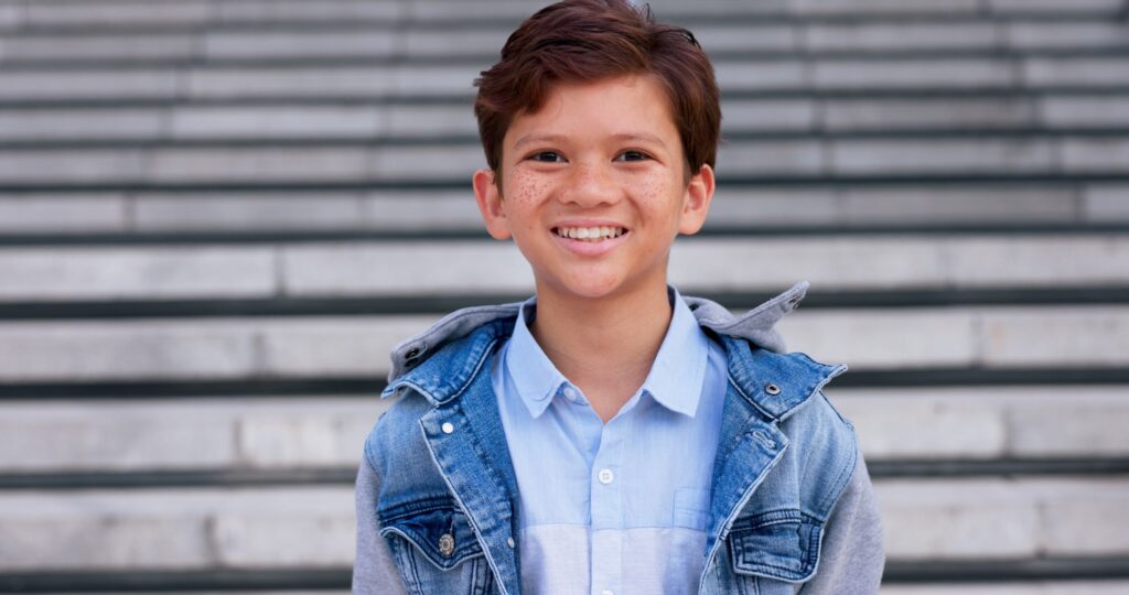 Niño con chaqueta vaquera y camisa azul claro sentado en unas escaleras al aire libre, representando la importancia de seguir precauciones y recomendaciones en el cuidado dental infantil.
