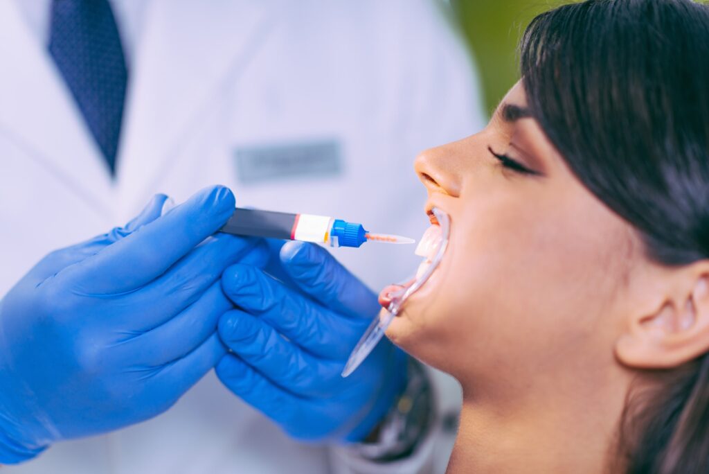 Dentista con guantes azules sosteniendo una jeringa o tubo de tratamiento dental junto al rostro de una paciente, ilustrando un procedimiento seguro y rápido realizado en un entorno clínico moderno.