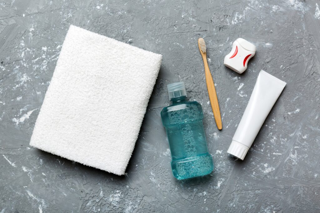 Productos de higiene bucal como colutorio, cepillo de dientes, hilo dental, pasta dental y toalla sobre una superficie gris, utilizados para ilustrar respuestas a preguntas frecuentes sobre el tratamiento con colutorios en la rutina diaria de salud oral.