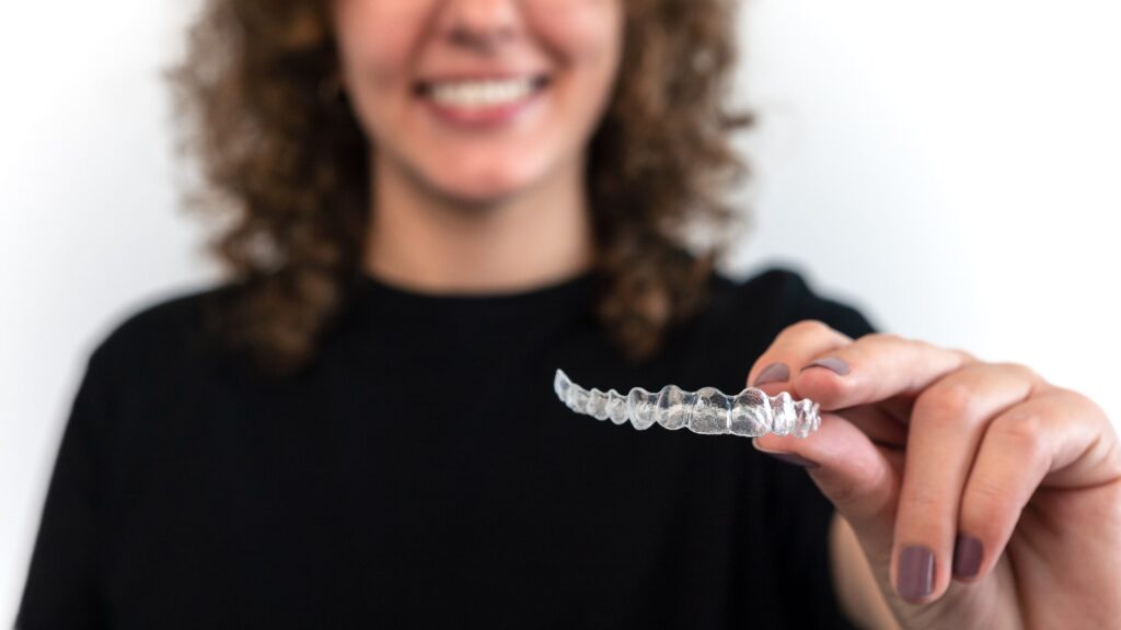 Mujer sonriendo sostiene un alineador transparente, ilustrando el proceso del tratamiento Invisalign para enderezar los dientes de forma discreta y cómoda.
